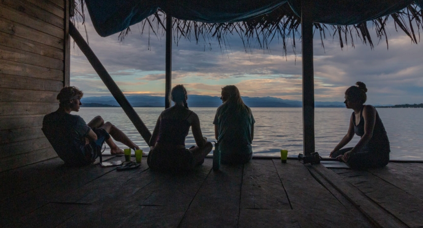 Four people sit on a dock with a thatched roof. The sky appears in gentle colors. 